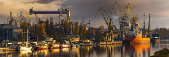 Panorama terenów przemysłowych nad Odrą. Widoczne są dźwigi i inne urządzenia portowe, budynki przemysłowe oraz nabrzeże ze statkami. 