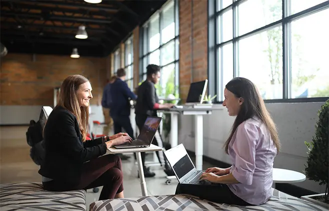 Two women are sitting across from each other in a modern office, talking while working on their laptops. Other employees can be seen in the background.
